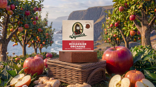 A Squatch Oceanside Orchard natural soap bar displayed on a wicker basket, surrounded by fresh red apples and sliced apples with ginger roots, set against a scenic background of an apple orchard and ocean cliffs.