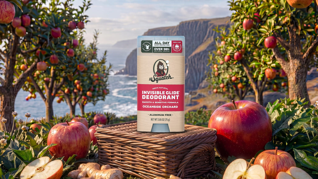 A package of Dr Squatch Invisible Glide Deodorant stands on a wicker basket in an orchard setting with apples and a coastal cliff in the background.