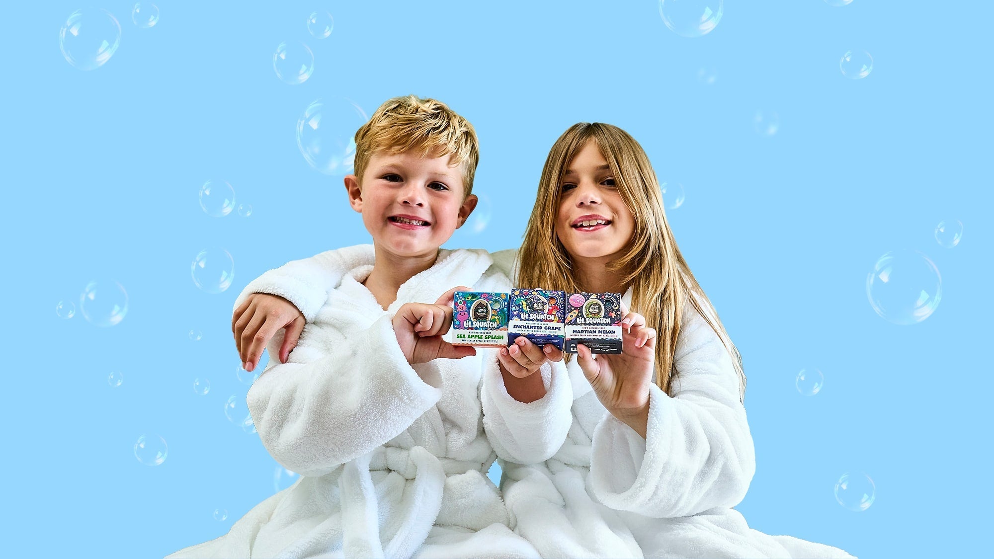 Two children in white robes holding three colorful Dr. Squatch soap boxes against a blue background with bubbles.
